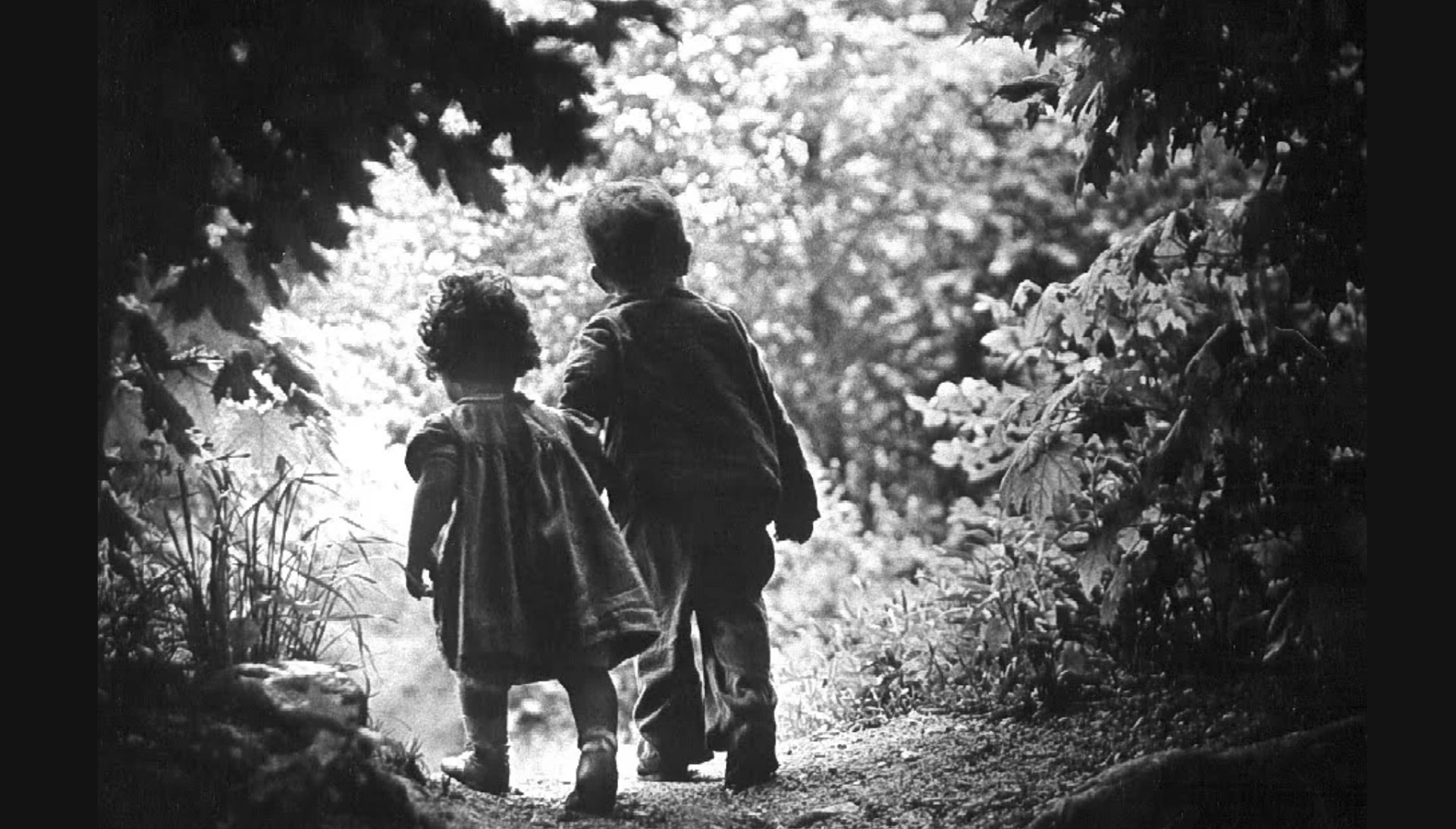 Two young children walking hand in hand toward a sunlit clearing in the woods, black and white photograph